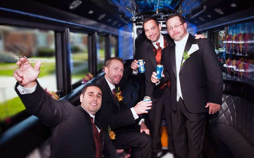 Groomsmen in dark suits holding beer cans inside a party bus with blue lights