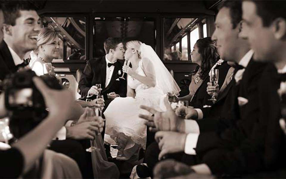 Bride and groom kissing inside a party bus as guests in tuxedos raise champagne
