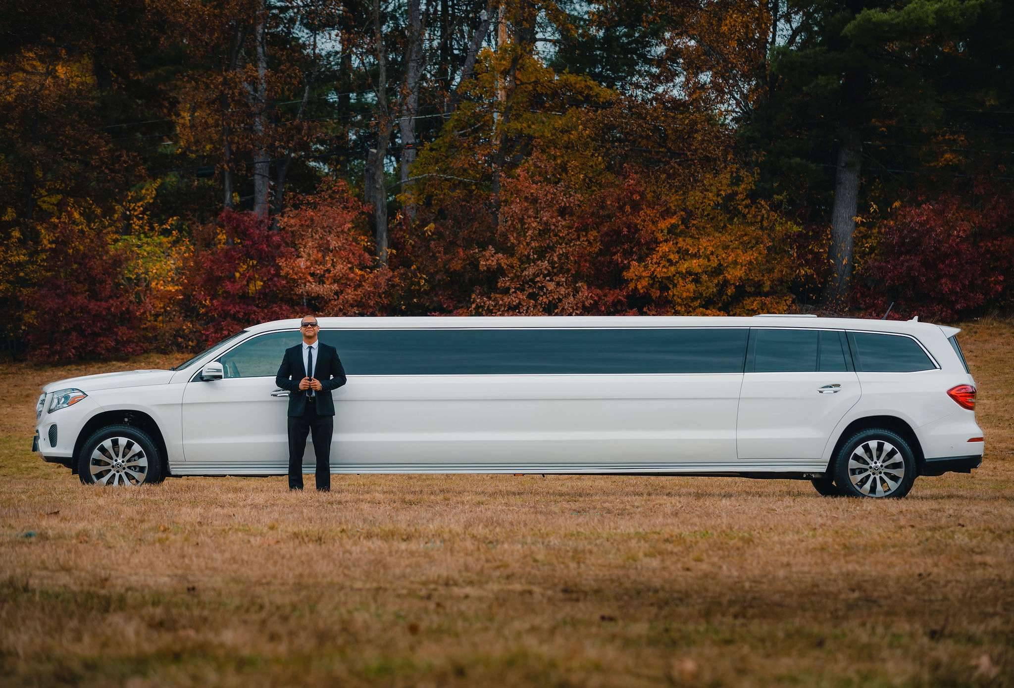 White Mercedes-Benz GLS stretch limousine with a chauffeur in a suit, autumn trees behind