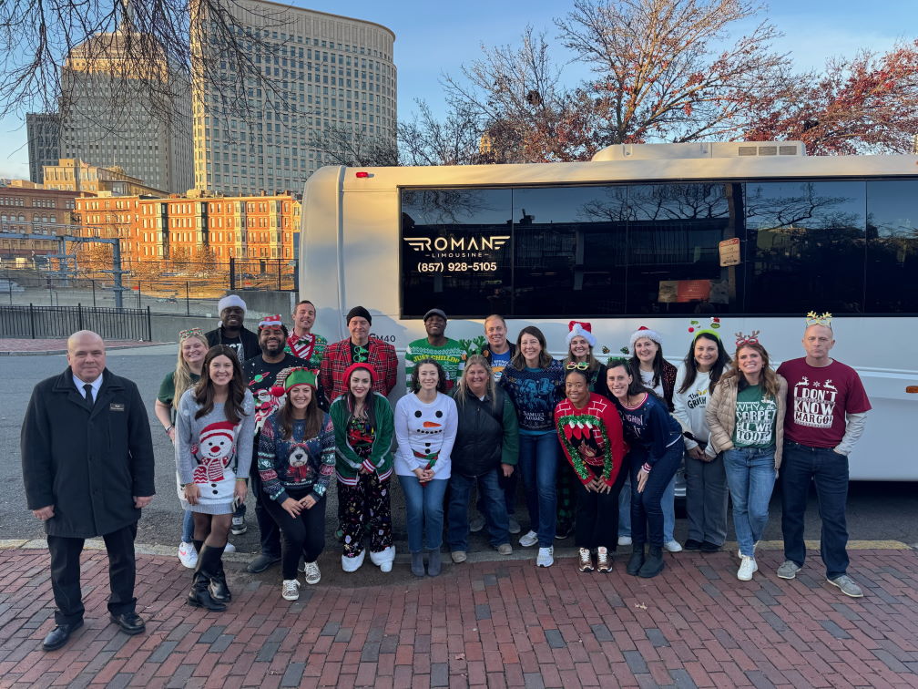 Group in holiday sweaters and Santa hats by a white Roman Limousine party bus