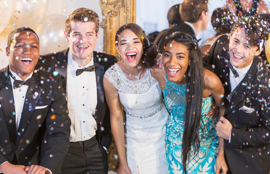 Young people in tuxedos and formal dresses laughing under falling confetti