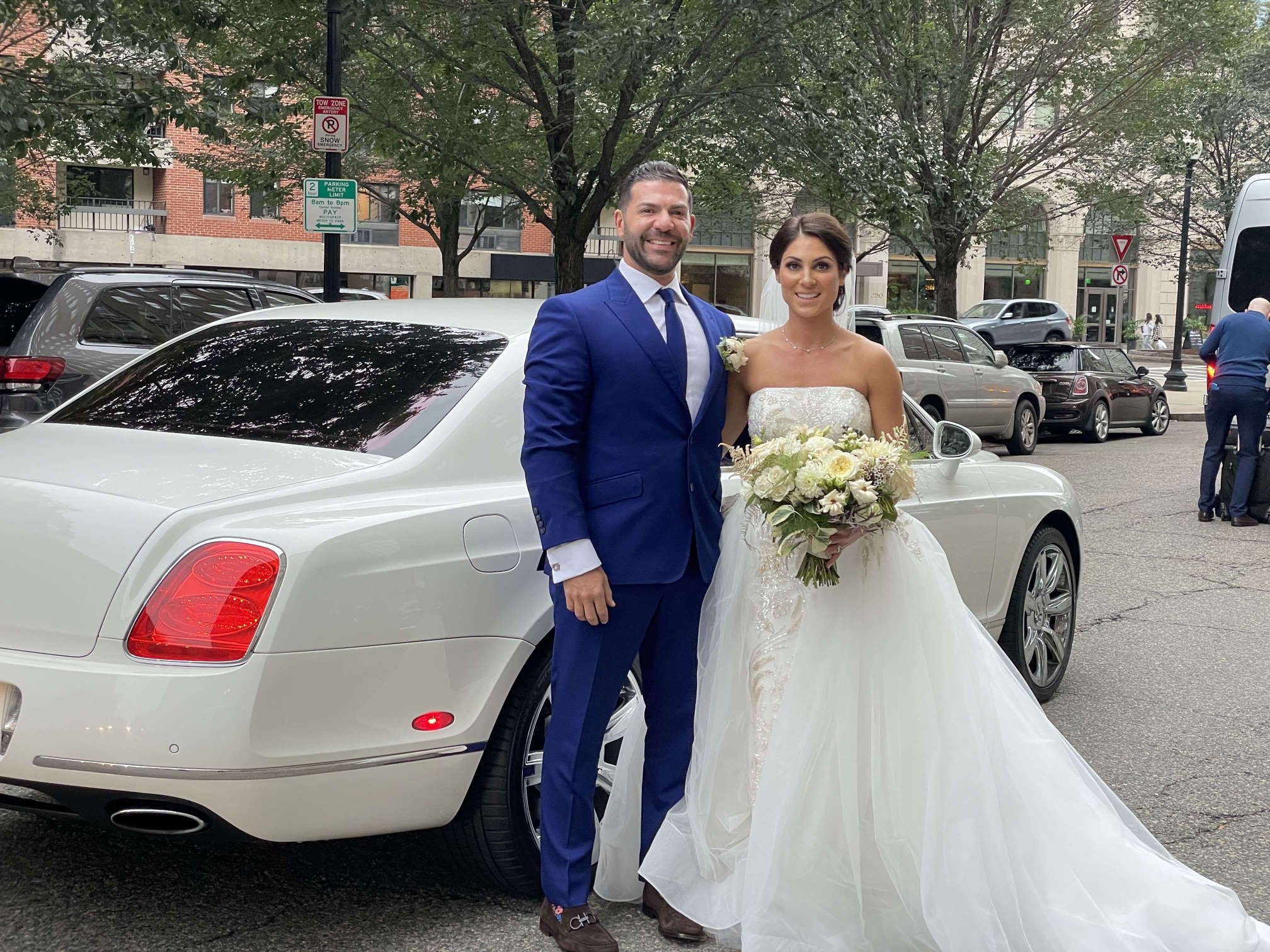 Bride and groom in a blue suit beside a white Bentley on a city street