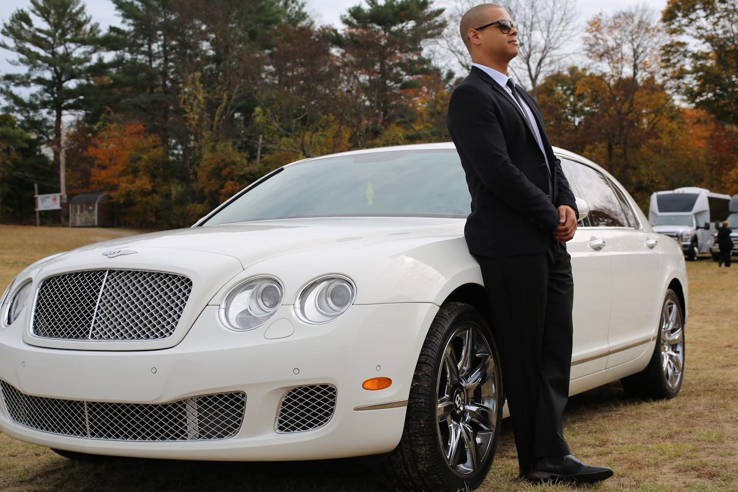 Chauffeur in a black suit leaning on a white Bentley sedan parked on grass