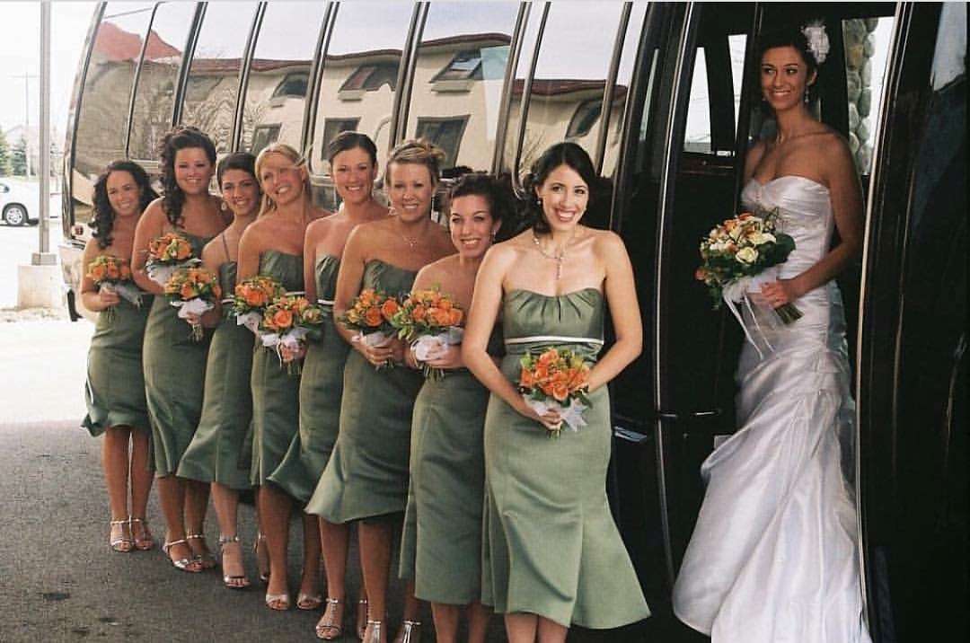 Bride and bridesmaids in green dresses with orange bouquets beside a black limo bus