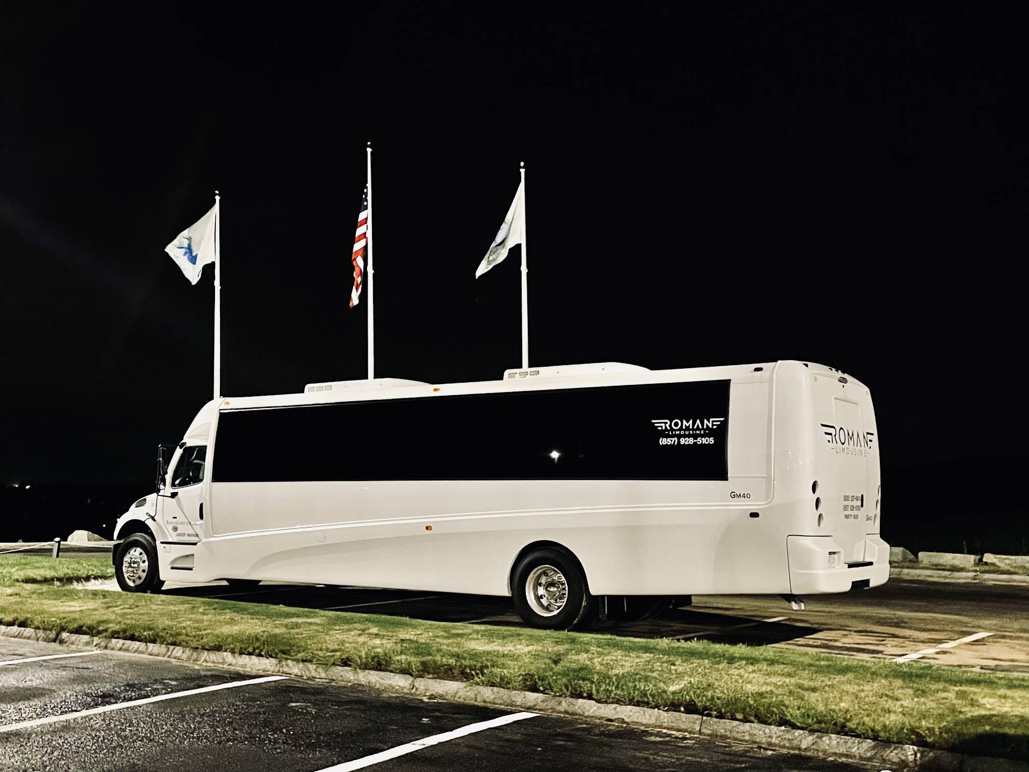 White party bus with Roman Limousine lettering parked at night beneath flagpoles