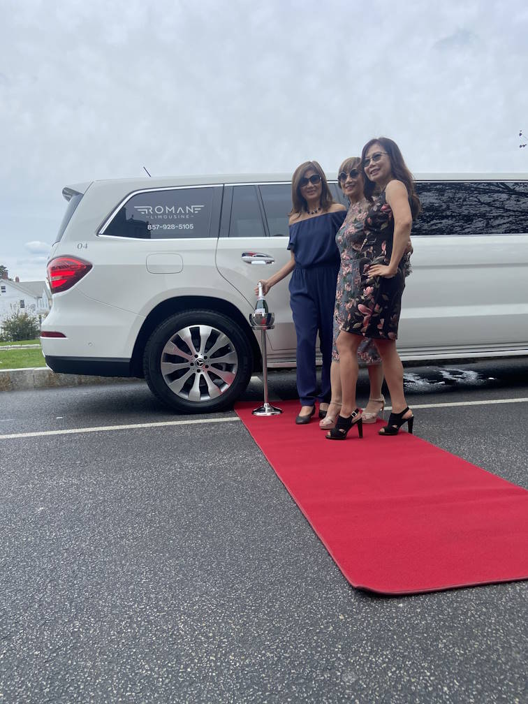 Three women on a red carpet beside a white Mercedes-Benz stretch limo lettered Roman Limousine