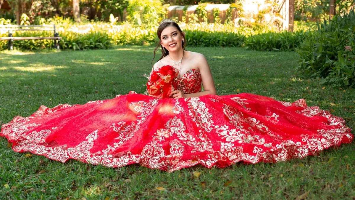 Girl in a red, gold-embroidered ball gown and tiara sitting on a lawn with roses