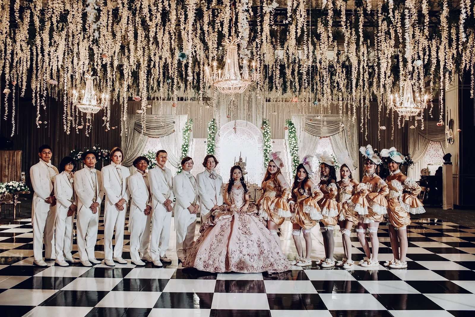 Girl in a pink ball gown with her court of friends in a chandelier-lit ballroom