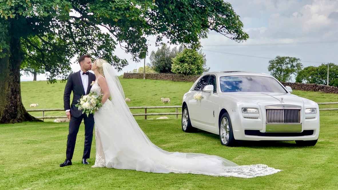 Bride and groom kissing on a lawn beside a white Rolls-Royce with flowers attached