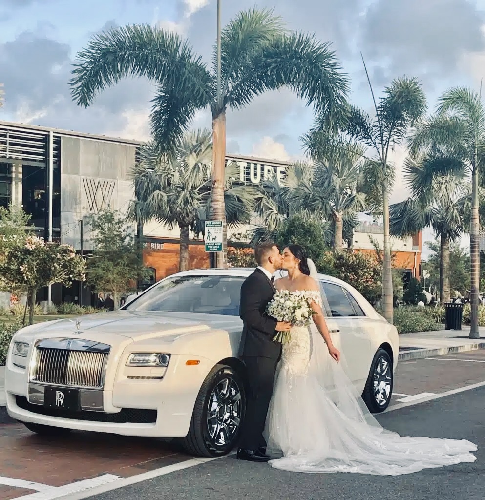 Bride and groom kissing beside a white Rolls-Royce Ghost under palm trees