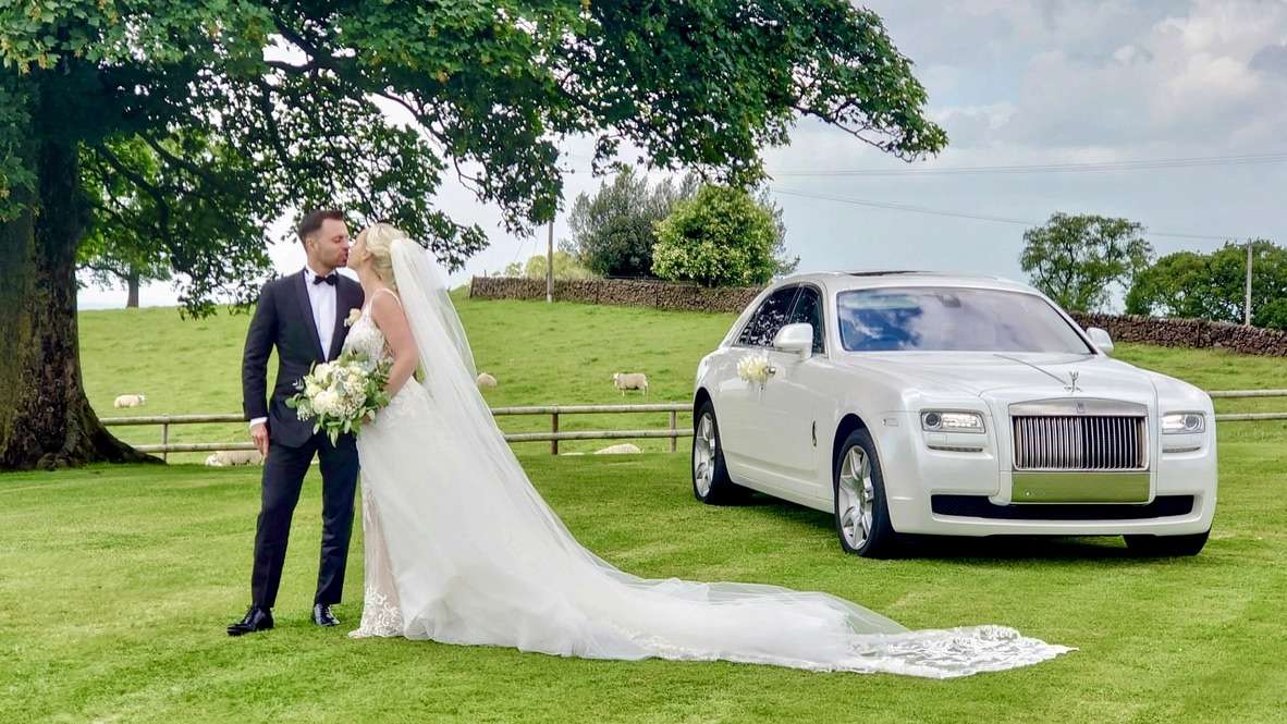 Bride and groom kissing on a lawn beside a white Rolls-Royce with flowers attached