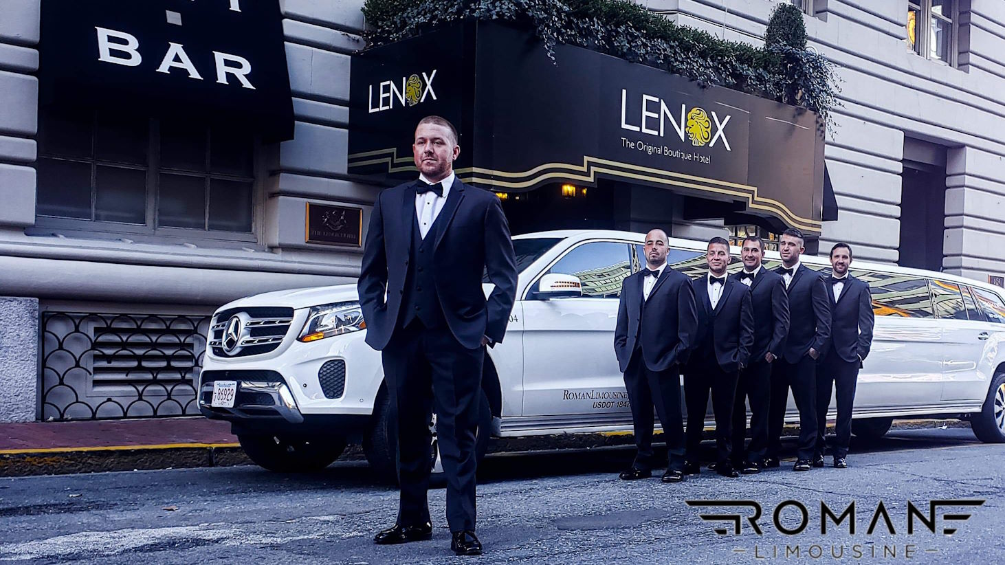 Groom and groomsmen in tuxedos beside a white Mercedes SUV limo outside the Lenox hotel