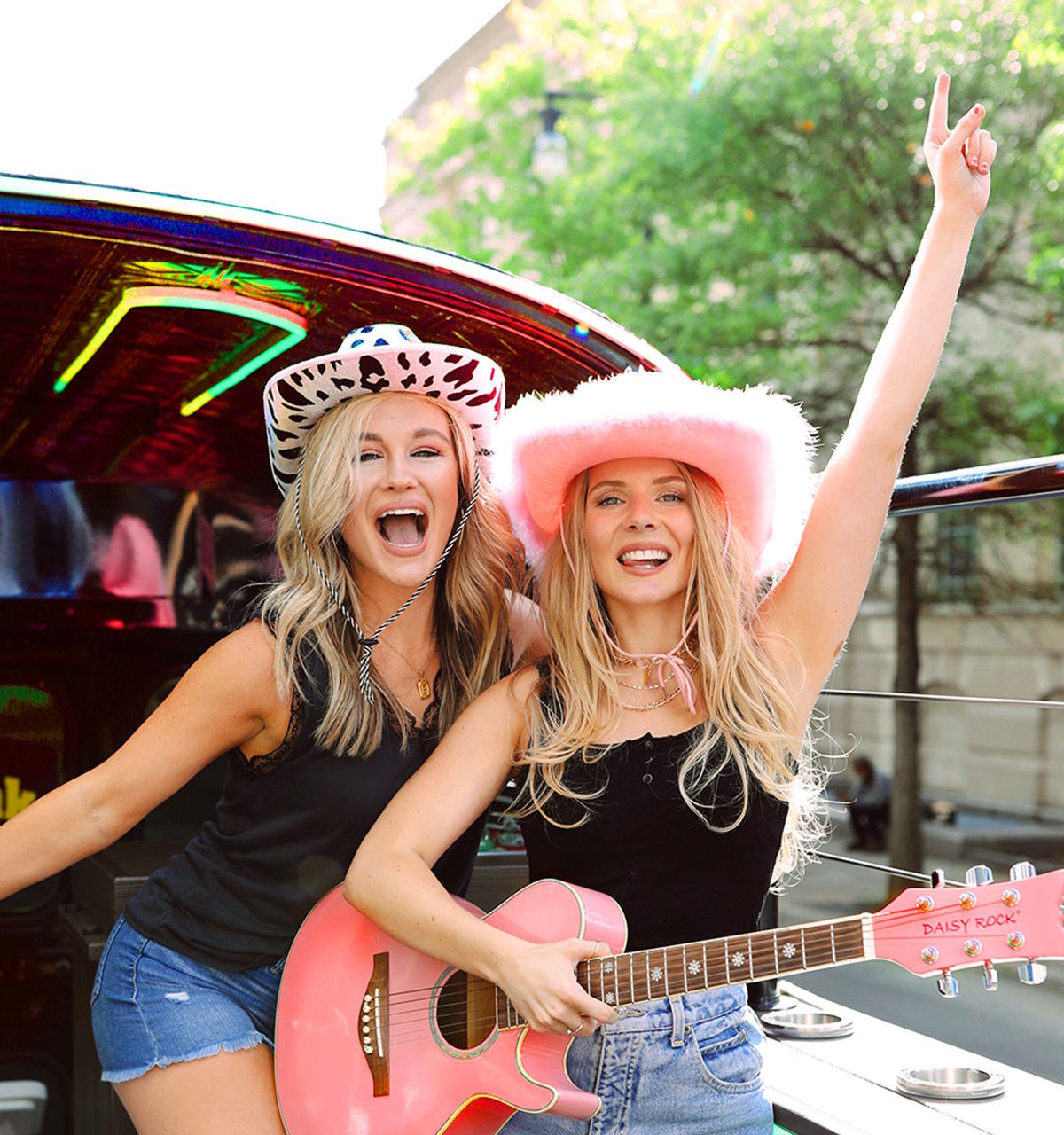 Two women in cowboy hats cheering by a neon-lit party vehicle, one holding a guitar