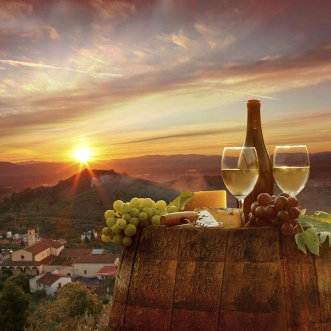 White wine bottle, two glasses, grapes and cheese on a barrel at sunset